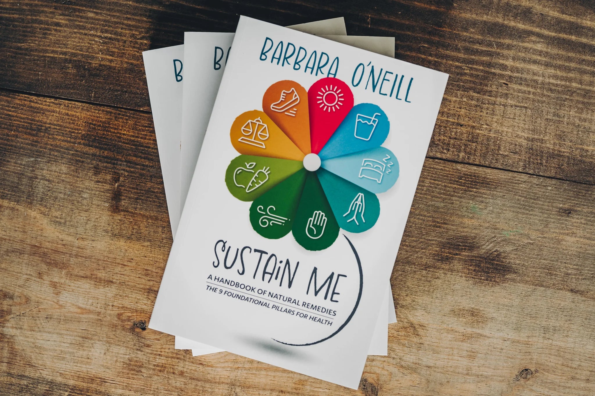 Stack of 'Sustain Me' books by Barbara O’Neill on rustic wooden table – natural health handbook featuring 9 foundational pillars of wellness and holistic remedies